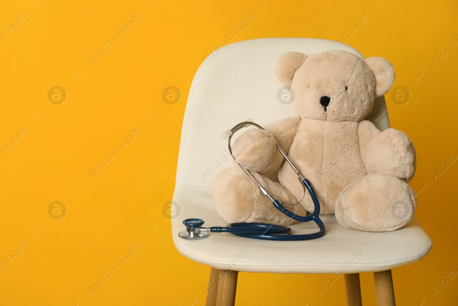Photo of Teddy bear with stethoscope on chair against yellow background, space for text. Pediatrician practice