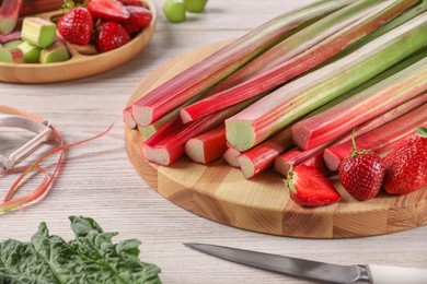 Photo of Cut fresh rhubarb stalks, strawberries and knife on white wooden table, closeup