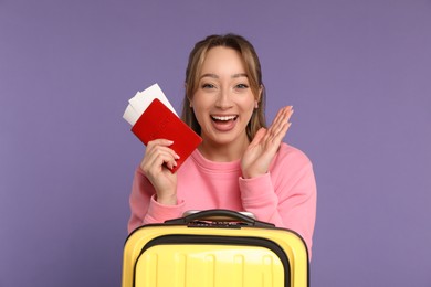 Happy young woman with passport, ticket and suitcase on purple background