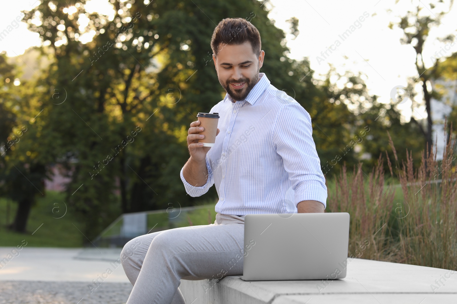Photo of Handsome young man with cup of coffee using laptop on stone bench outdoors