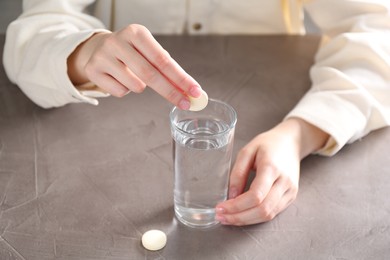 Woman putting effervescent pill into glass of water at grey table, closeup