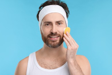 Man with headband washing his face using sponge on light blue background