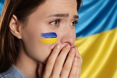 Sad young woman with clasped hands near Ukrainian flag, closeup