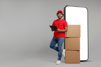Courier with stack of parcels and clipboard near huge smartphone on grey background. Delivery service. Space for text