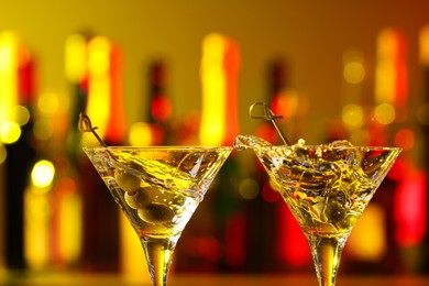 Photo of Clinking martini glasses with splashing cocktail and olives in bar, closeup