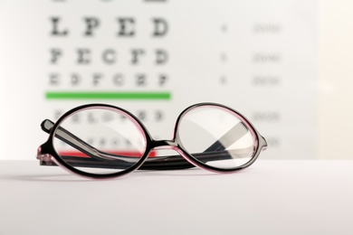 Photo of Glasses with corrective lenses on table against eye chart