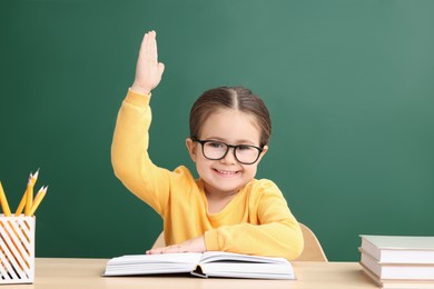 Photo of Happy little school child raising hand while sitting at desk with books near chalkboard