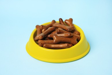 Photo of Yellow bowl with bone shaped dog cookies on light blue background, closeup
