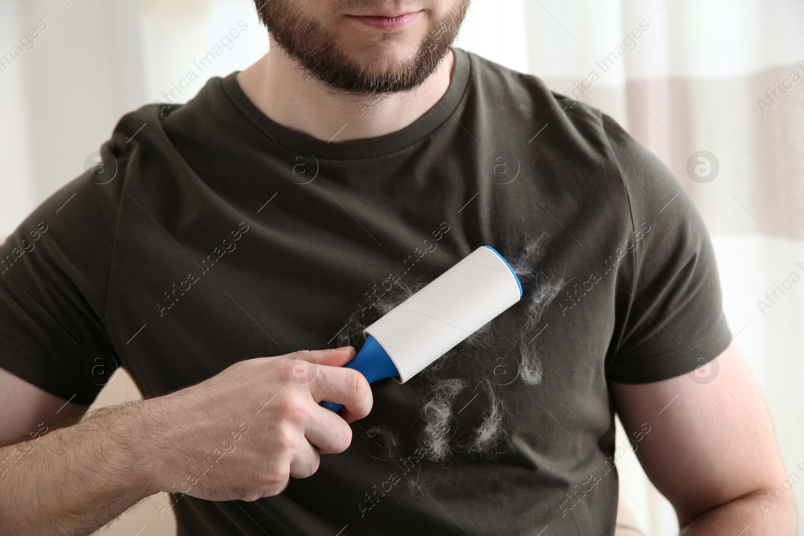 Photo of Man removing hair from grey t-shirt with lint roller on light background, closeup