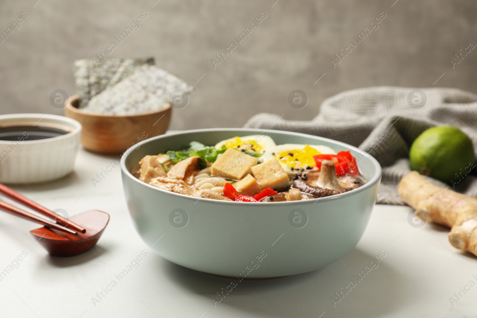 Photo of Bowl of delicious ramen, ingredients and chopsticks on white table, closeup. Noodle soup