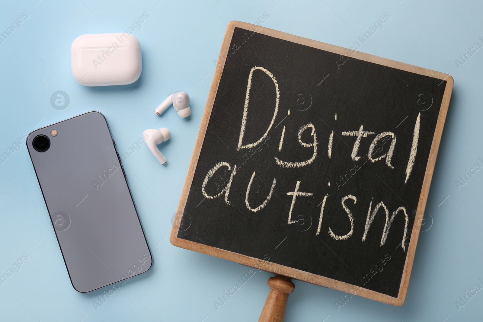 Photo of Small chalkboard with written phrase Digital Autism, smartphone and earphones on light blue background, flat lay