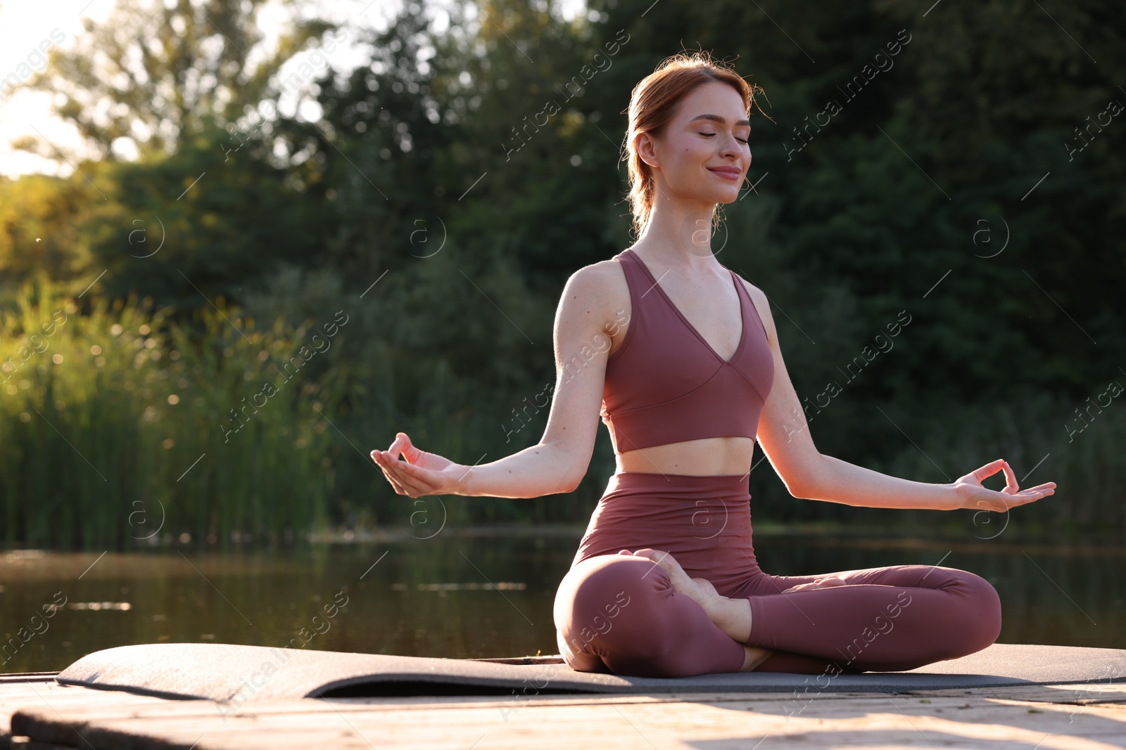 Photo of Beautiful woman practicing Padmasana on yoga mat on wooden pier near pond, space for text. Lotus pose