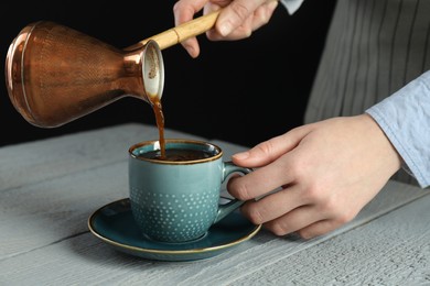 Turkish coffee. Woman pouring brewed beverage from cezve into cup at gray wooden table against black background, closeup