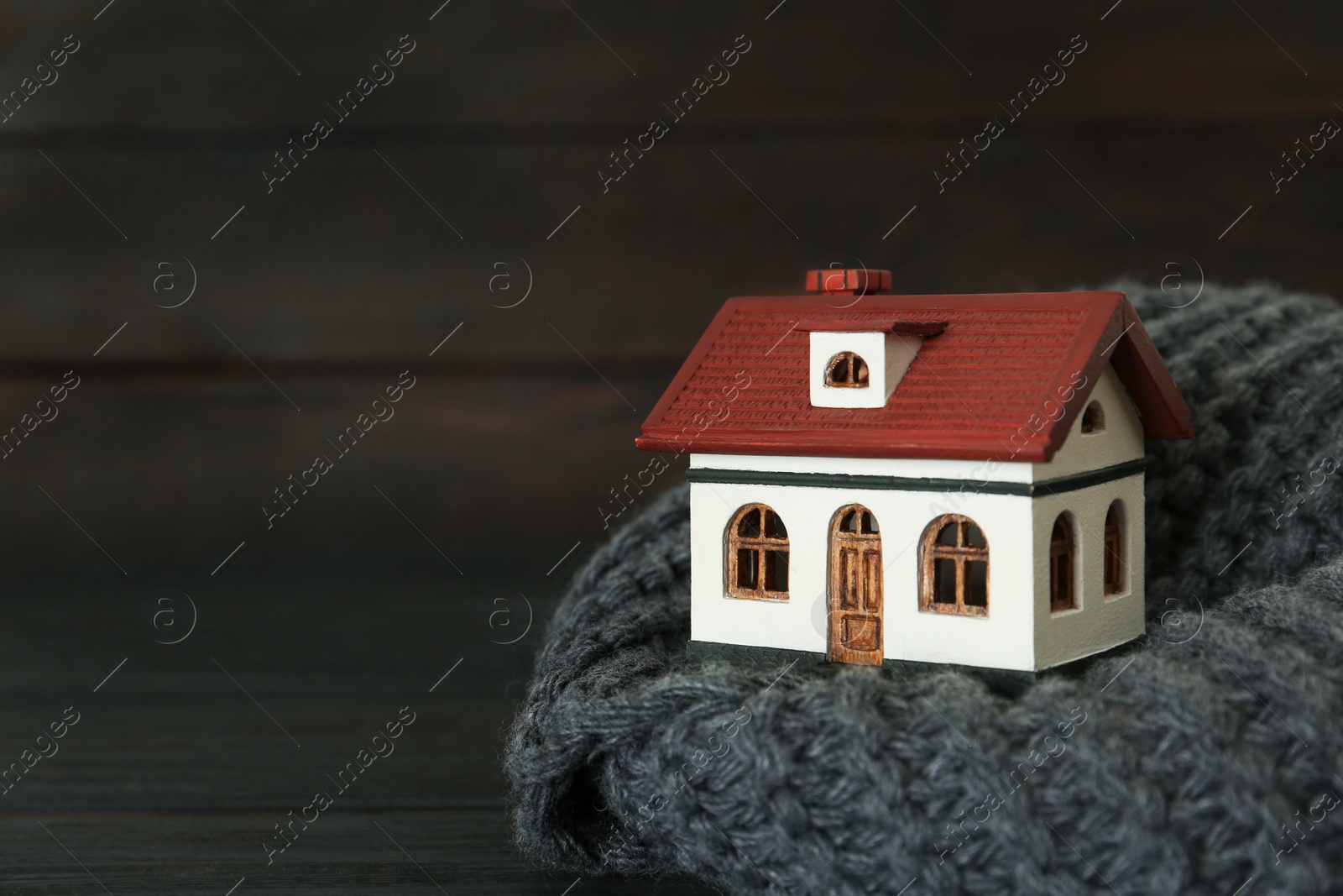 Photo of House model and scarf on black table against wooden background, space for text. Heating efficiency