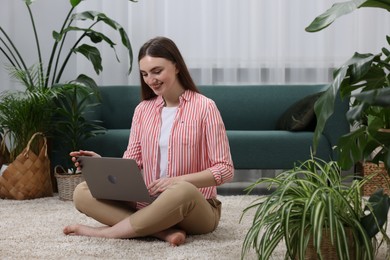 Beautiful young woman using laptop on floor in room with green houseplants