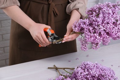Photo of Woman trimming lilac branches with secateurs at white wooden table, closeup