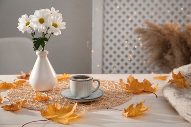 Photo of Cup of aromatic coffee, beautiful flowers and autumn leaves on white wooden table indoors, space for text
