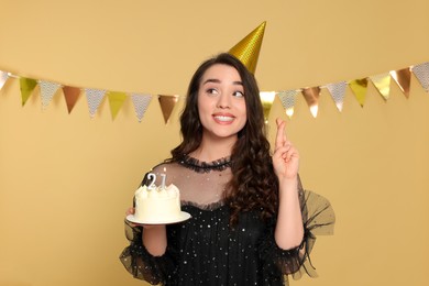 Photo of Coming of age party - 21st birthday. Smiling woman holding delicious cake with number shaped candles and crossing fingers on beige background