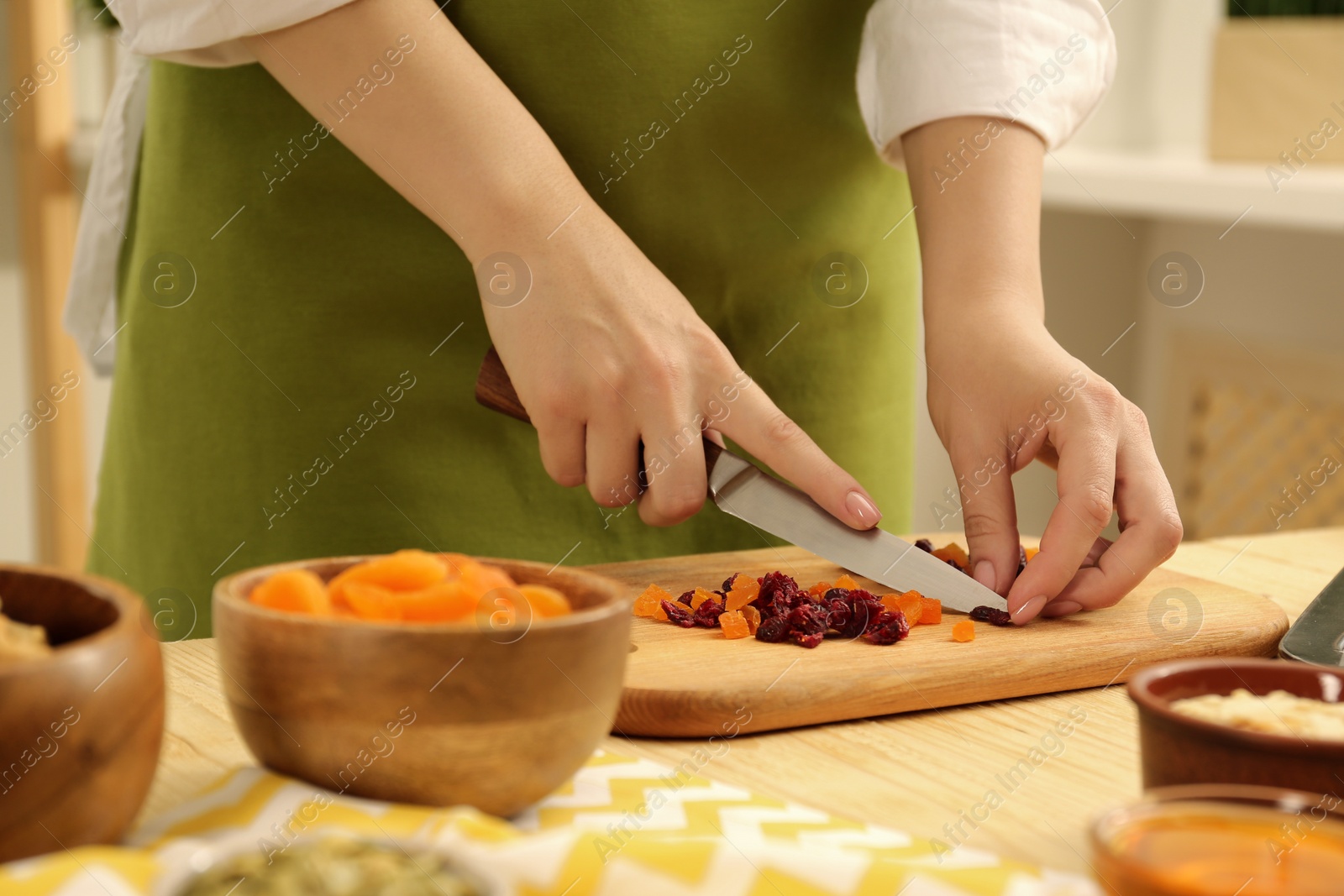 Photo of Making granola. Woman cutting dried apricots and cherries at table in kitchen, closeup