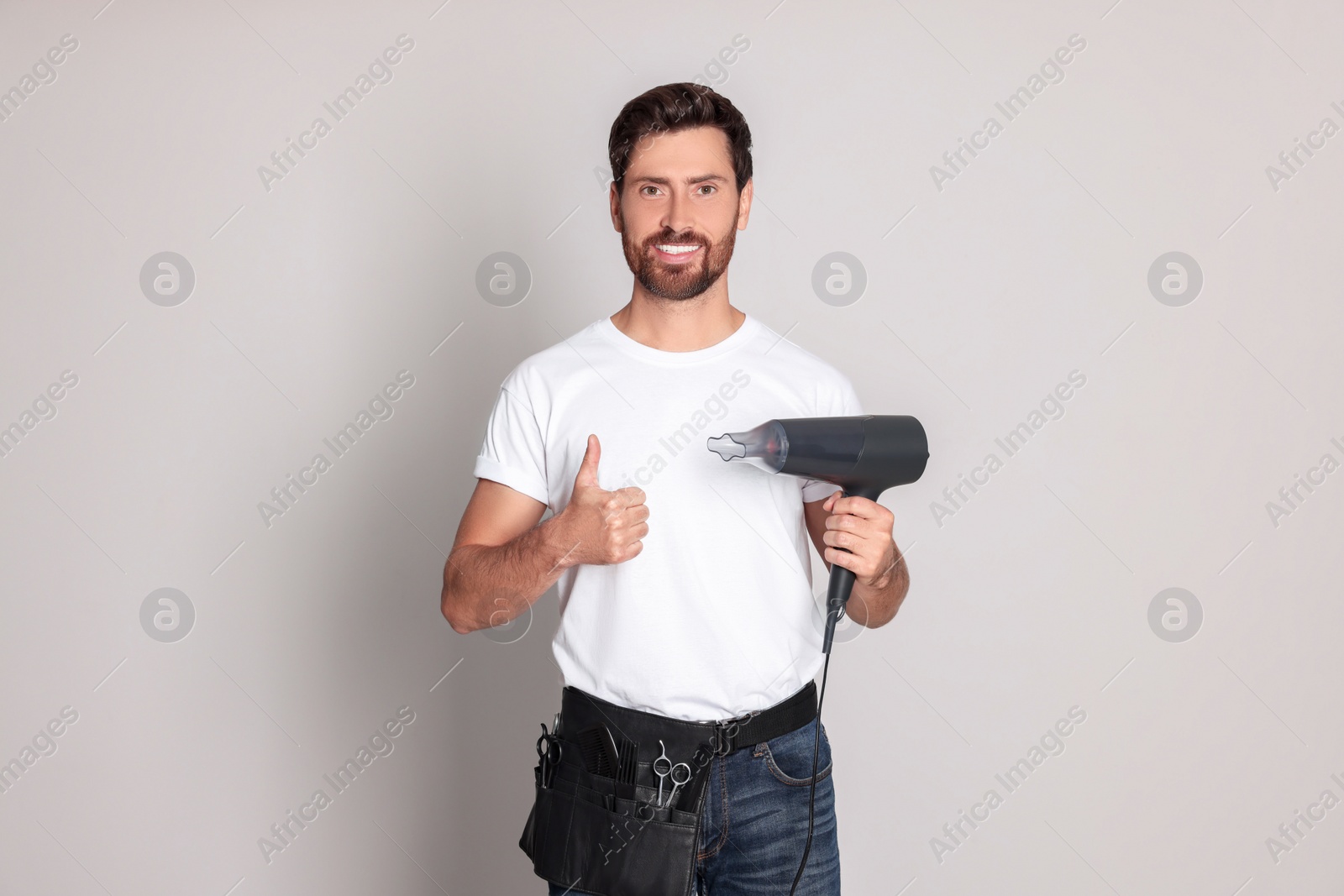 Photo of Smiling hairdresser with tool bag showing thumb up holding dryer on light grey background