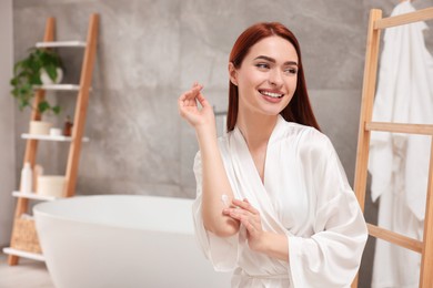 Beautiful young woman applying body cream onto elbow in bathroom, space for text