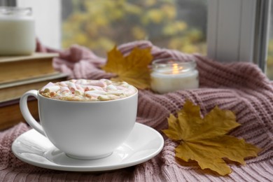 Photo of Cup of tasty hot drink with marshmallows near books, leaves and burning candle on blanket, closeup. Autumn coziness