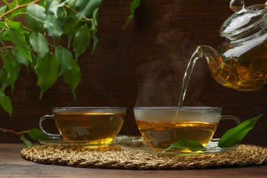 Pouring green tea into glass cup with saucer and leaves on wooden table