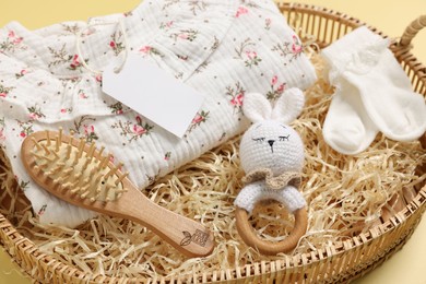 Different baby accessories, clothes and blank card in wicker basket on yellow background, closeup