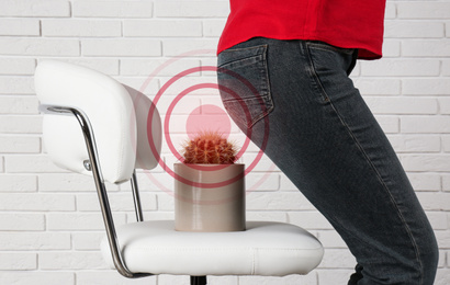 Image of Hemorrhoid concept. Woman sitting down on chair with cactus near white brick wall, closeup