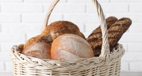 Different types of bread in wicker basket against white brick wall, closeup