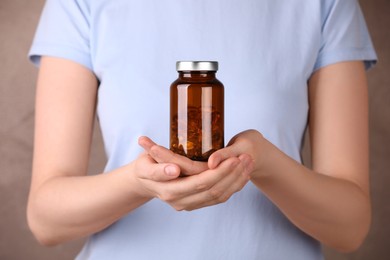 Photo of Woman holding bottle with vitamin capsules against light brown background, closeup