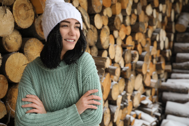 Photo of Young woman wearing warm sweater and hat near stack of firewood outdoors, space for text. Winter season