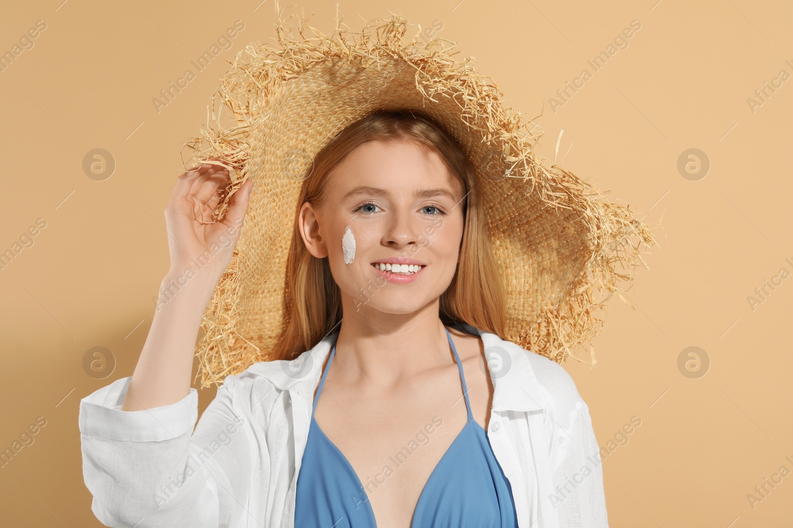 Photo of Beautiful young woman in straw hat with sun protection cream on her face against beige background