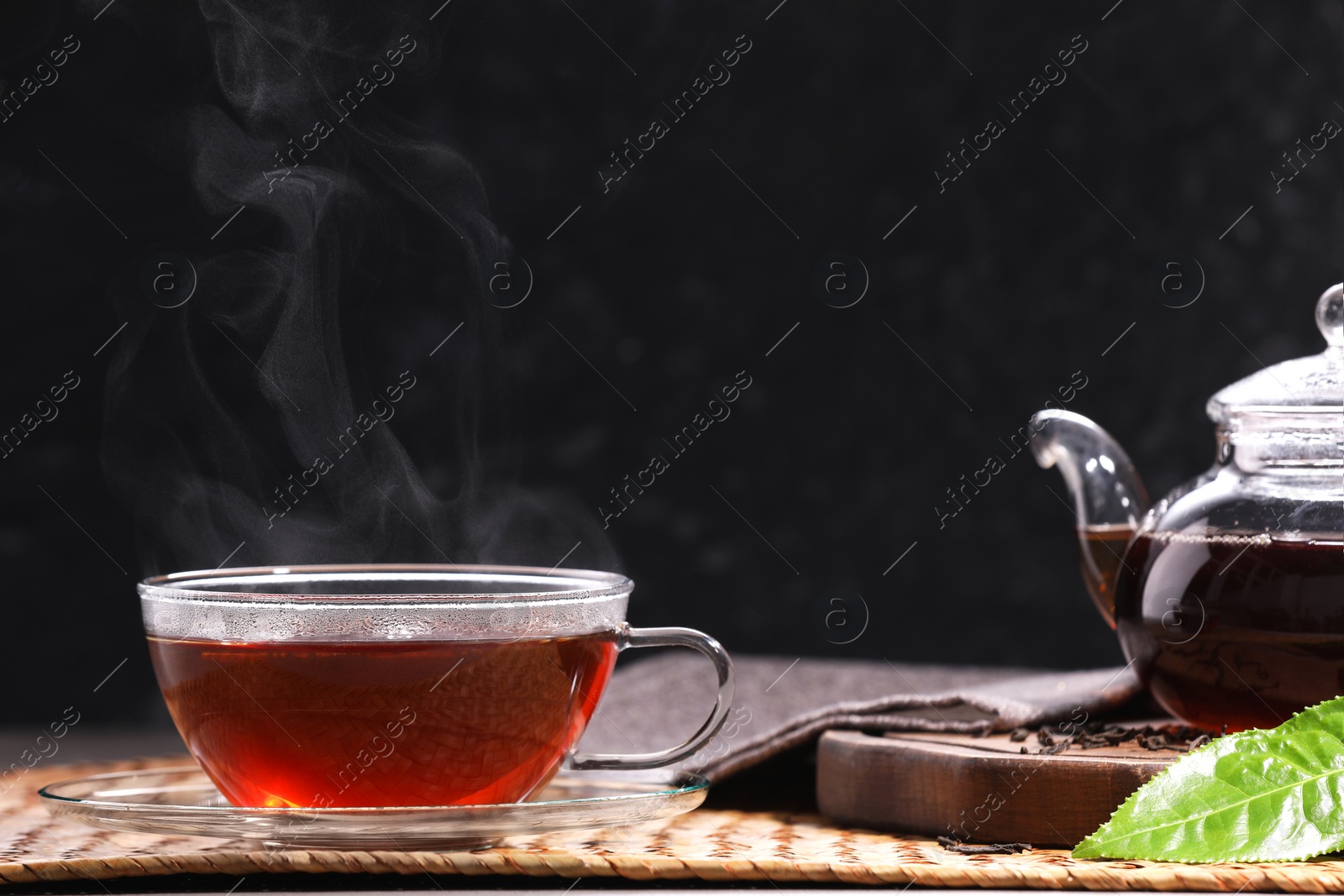 Photo of Aromatic hot tea in glass cup with saucer, teapot and leaves on table against black background. Space for text