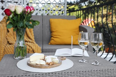 Vase with roses, open book, glasses of wine and snacks on rattan table at balcony