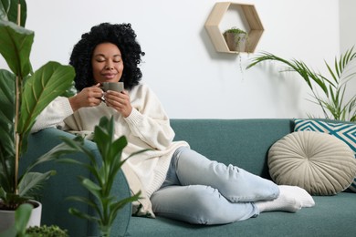 Relaxing atmosphere. Woman with cup of hot drink on sofa near beautiful houseplants in room