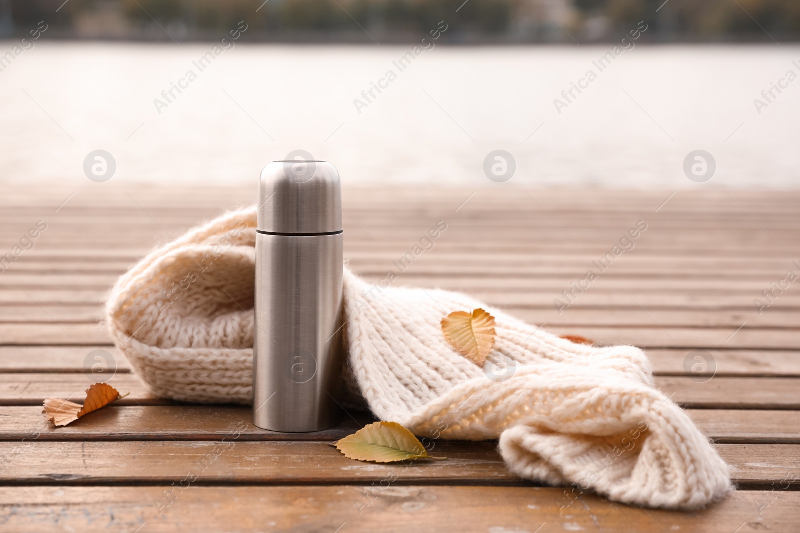 Photo of Metallic thermos, knitted scarf and fallen leaves on wooden pier near river. Space for text