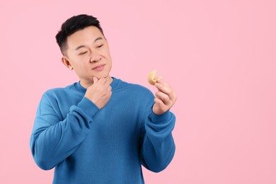 Asian man holding tasty fortune cookie with prediction on pink background. Space for text