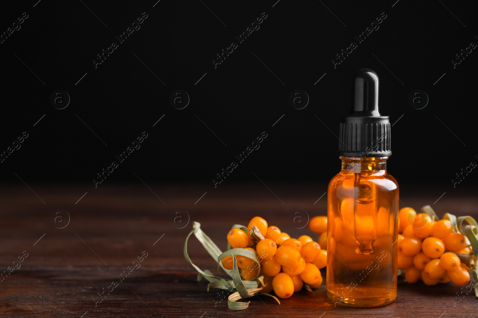 Photo of Ripe sea buckthorn and bottle of essential oil on wooden table against black background. Space for text