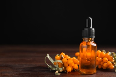 Photo of Ripe sea buckthorn and bottle of essential oil on wooden table against black background. Space for text