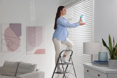 Photo of Woman on metal ladder wiping blinds at home