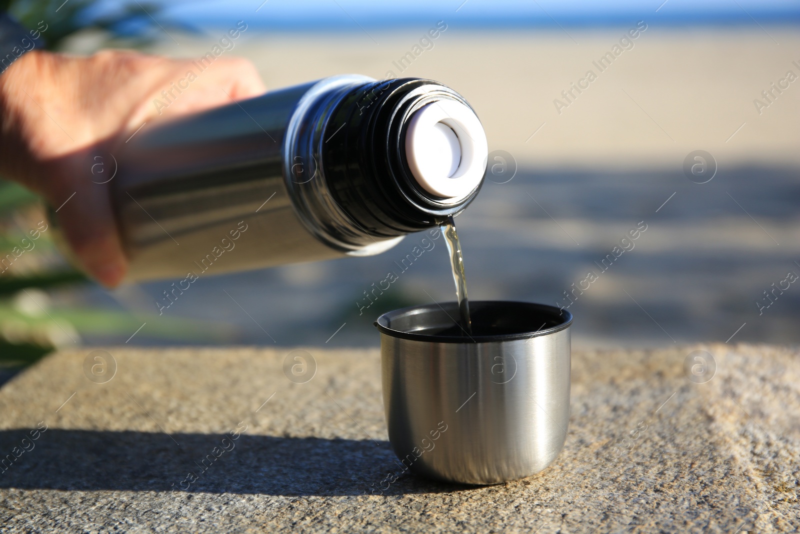 Photo of Woman pouring hot drink from metallic thermos into cap on stone surface outdoors, closeup