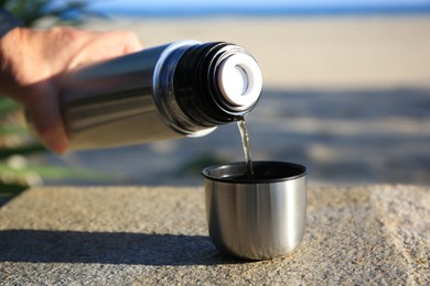 Woman pouring hot drink from metallic thermos into cap on stone surface outdoors, closeup