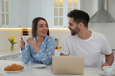 Photo of Happy couple in pajamas with laptop having breakfast at kitchen table