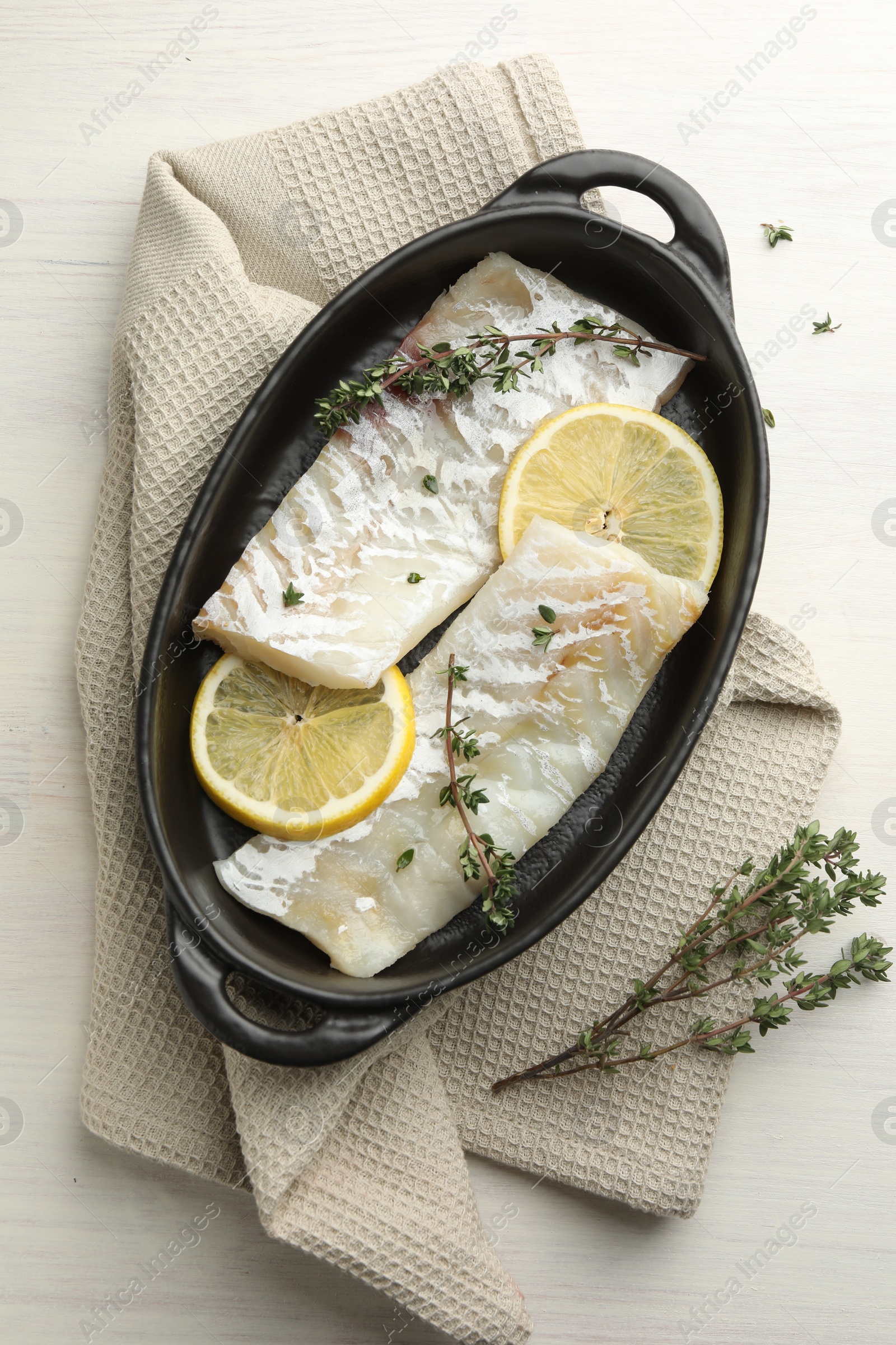 Photo of Fresh raw cod fillets with thyme and lemon in baking dish on white wooden table, top view