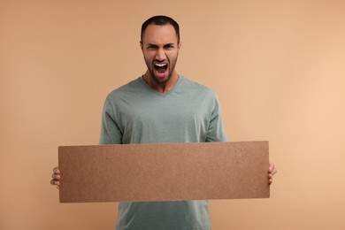 Angry man holding blank cardboard banner on beige background, space for text