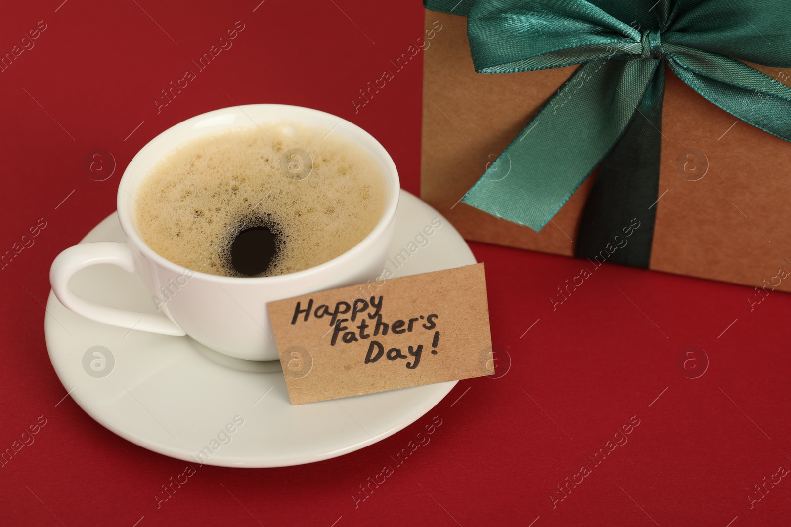 Photo of Happy Father's Day. Coffee cup, card with greetings and gift box on red table, closeup