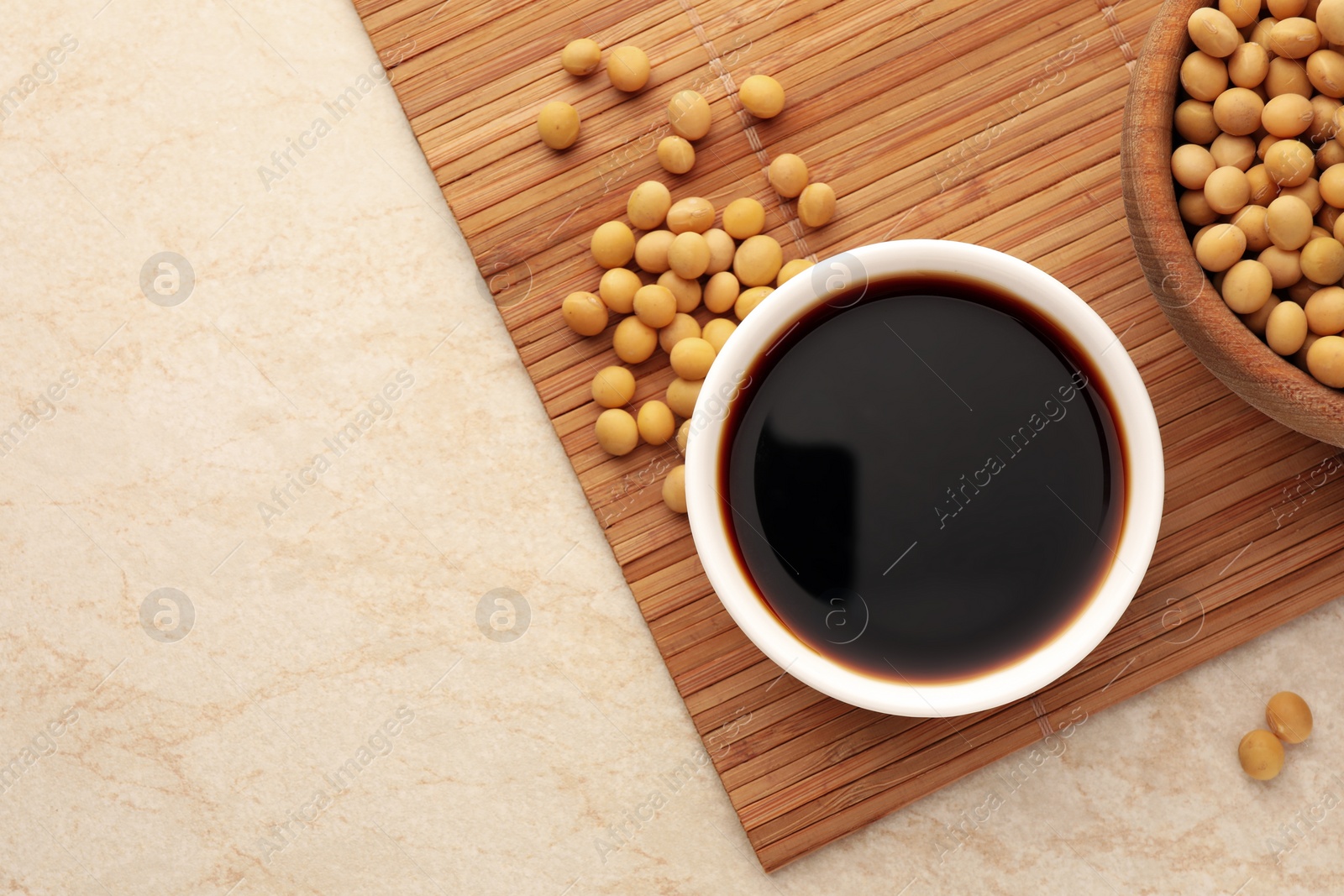 Photo of Soy sauce in bowl and soybeans on beige table, flat lay. Space for text