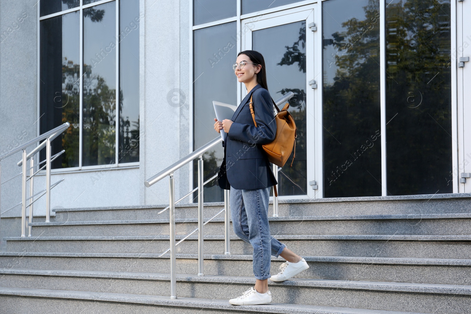 Photo of Beautiful young woman with stylish backpack and laptop near building outdoors