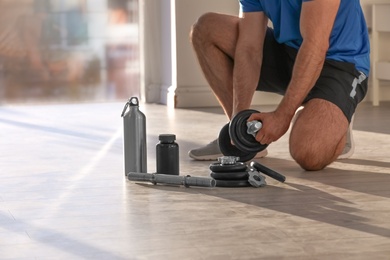 Photo of Male athlete assembling adjustable dumbbell near bottle, parts and jar of protein on floor indoors